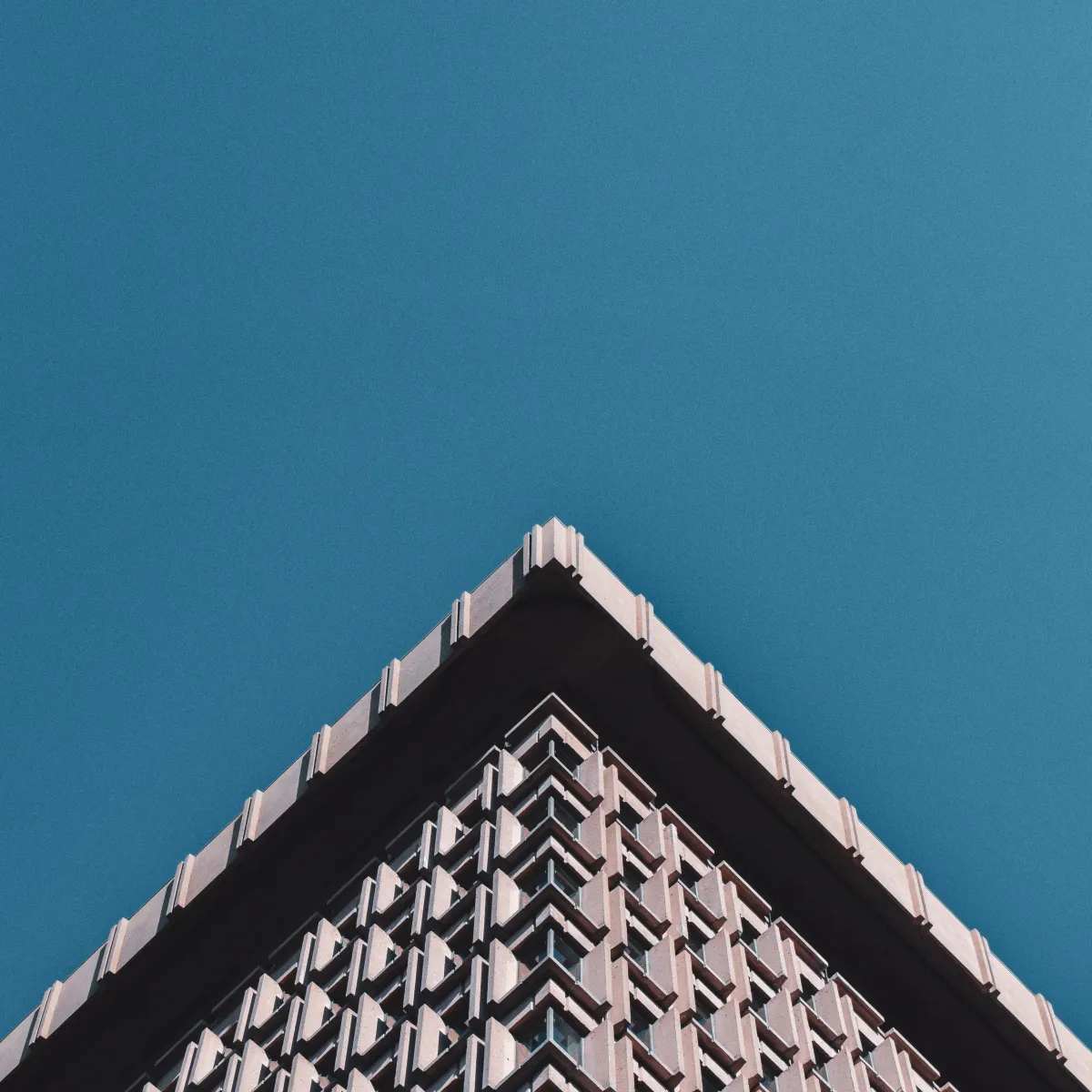 En vue de contre-plongée, un bâtiment d'architecture brutaliste comporte le quart inférieur de l'image, formant une pointe triangulaire, sur fond de ciel bleu immaculé.