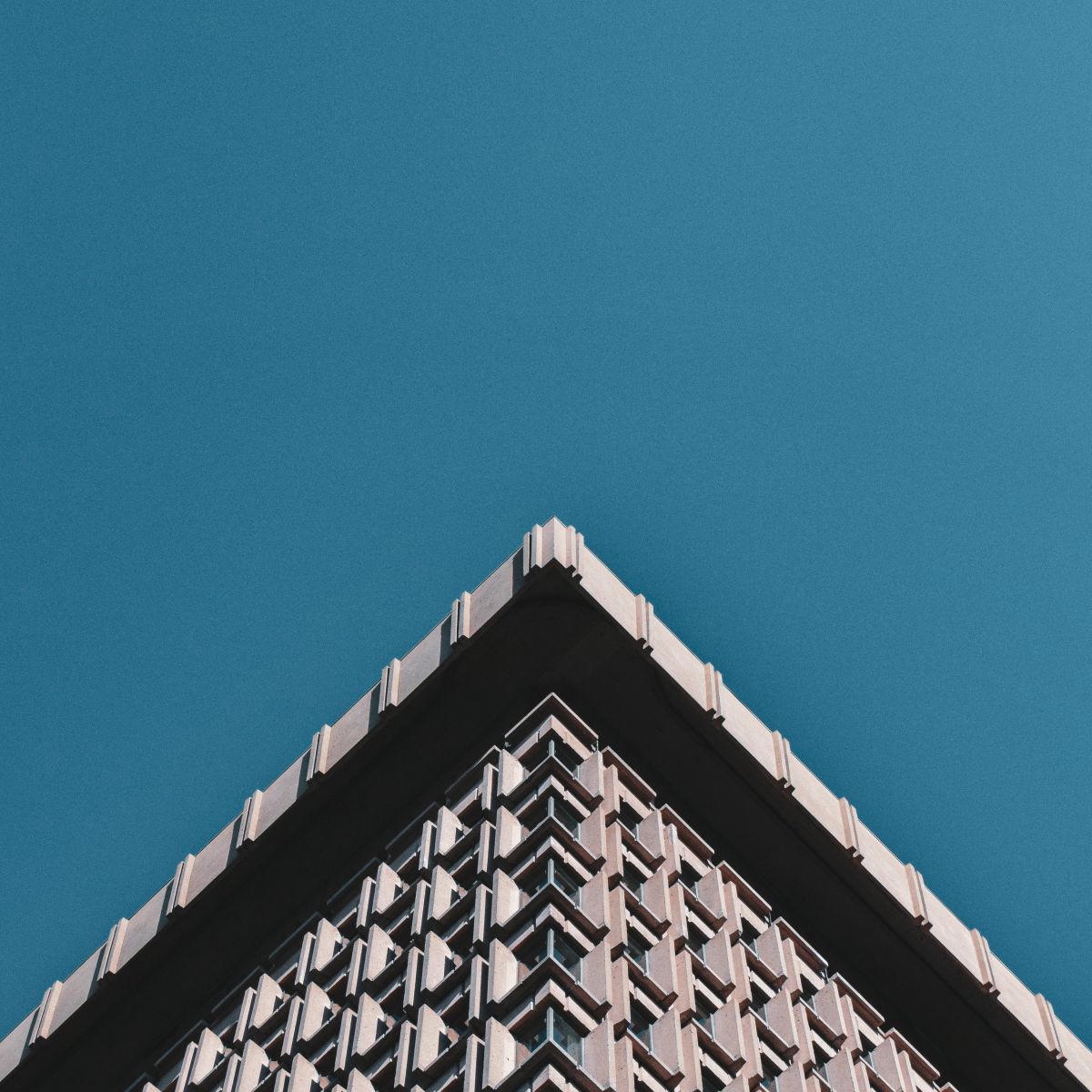 En vue de contre-plongée, un bâtiment d'architecture brutaliste comporte le quart inférieur de l'image, formant une pointe triangulaire, sur fond de ciel bleu immaculé.