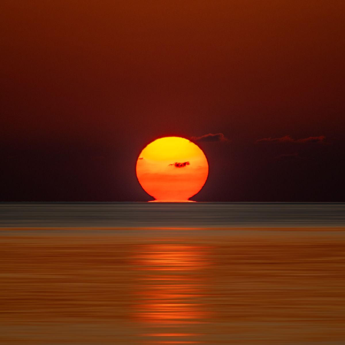 Un soleil rougeoyant se couchant sur l'océan, touchant l'horizon. Quelques petits nuages sont présents. Le ciel est mat et écarlate.
