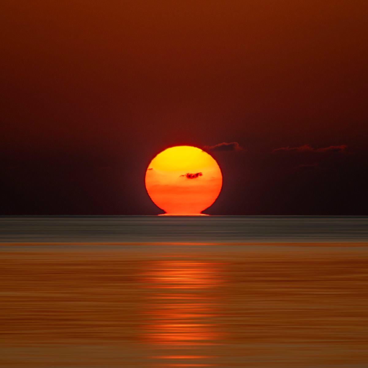 Un soleil rougeoyant se couchant sur l'océan, touchant l'horizon. Quelques petits nuages sont présents. Le ciel est mat et écarlate.